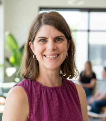 Dr. Kathe Pelletier Headshot of a woman with shoulder length dark brown hair wearing a purple dress in front of a blurry background where people are gathering.